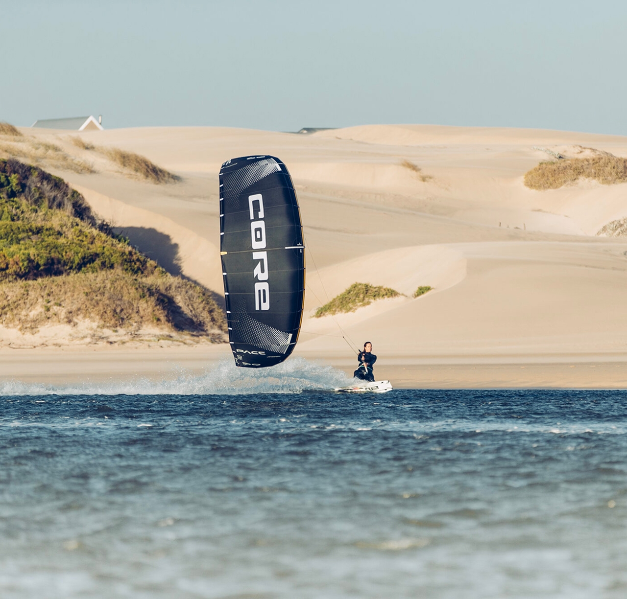 Praticante pilotando kite Core preto entre dunas de areia, casa ao fundo